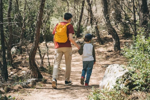 Rear view of tourists school boy and his dad walking a stone footpath in spring forest. Child kid and father wearing casual clothes and yellow backpack while hiking in summer greenwood leaf forest.