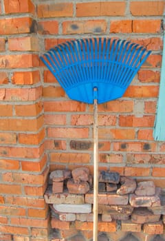 Blue plastic rake for harvesting leaves and grass are near the red brick wall.