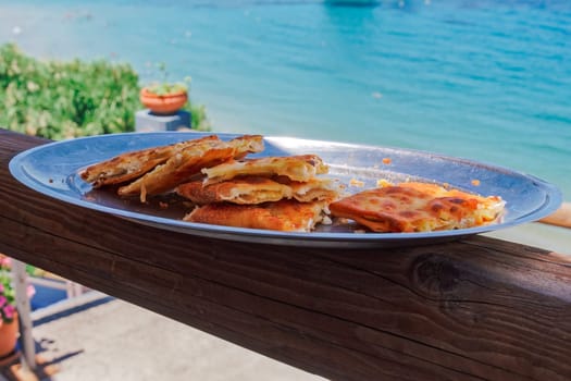 A dish with Greek tyropita, feta cheese pie portions with layers of buttered phyllo pastry against a defocused seafront background.