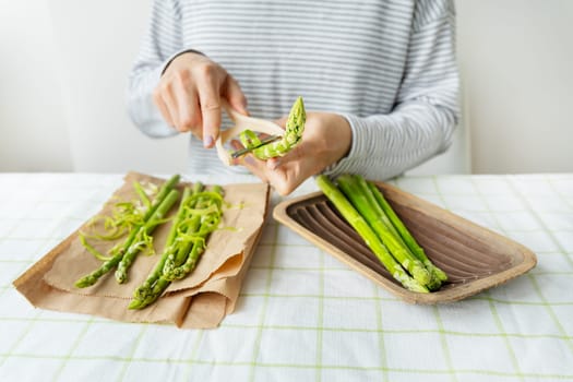 A woman cleans asparagus with a knife at the table and puts it in a wooden plate. Clean and wholesome food for health. Vegetarianism, raw food