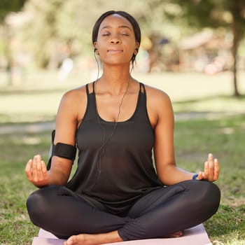 Yoga, black woman meditation in park with lotus pose, earphone for mindfulness podcast and zen outdoor in nature. Peace, spiritual energy and balance self care and music with fitness and wellness.