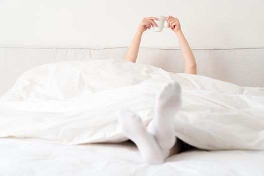 Female hand holding cup of coffee from under a blanket in bed. Woman waking up in the morning.