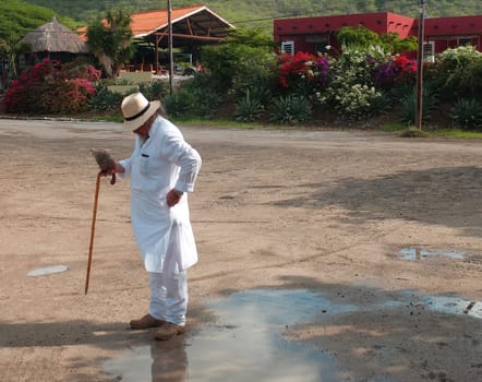 A tourist in white clothes (kurta), a stick and a straw hat standing in front of a row houses. He is checking his pocket. Caribbean vacation.