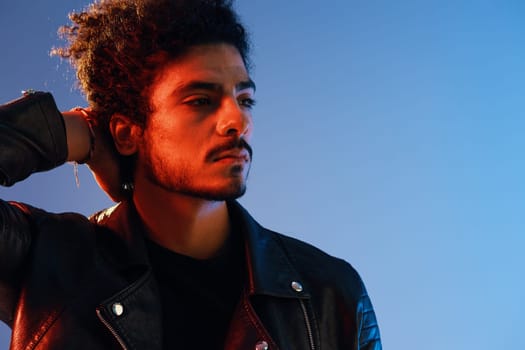 Portrait of a stylish man with curly hair on a blue background multinational, colored light, black leather jacket trend, modern concept. High quality photo