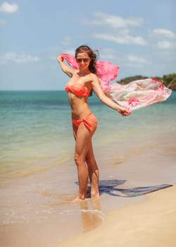 Young woman wearing bikini standing on wet sand at the beach, holding scarf waving in wind. Clear sea and sky background