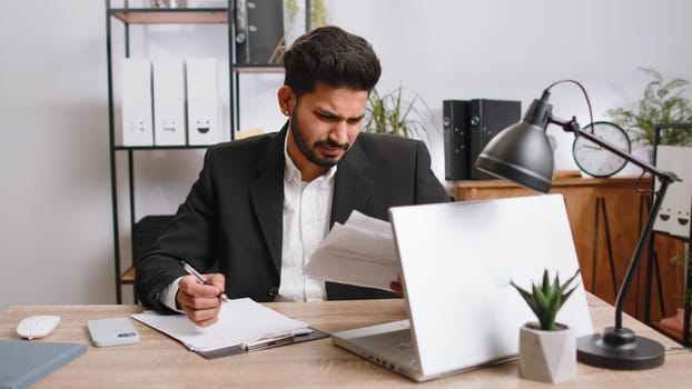 Stressed indian businessman working looking at unpaid bank debt bills, doing paperwork, planning budget, calculate finances mortgage payments at office. Displeased freelancer man throws paper bills