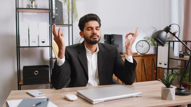 Indian businessman working on laptop computer, meditating, doing yoga breathing exercise in lotus position at home office. Calm serene freelancer arab man taking break. Busy occupation. Peace of mind