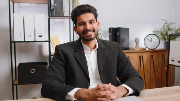 Portrait of happy smiling bearded indian businessman at modern home office workplace desk looking at camera. Male hindu in formal suit. Handsome man remote distant online working on laptop