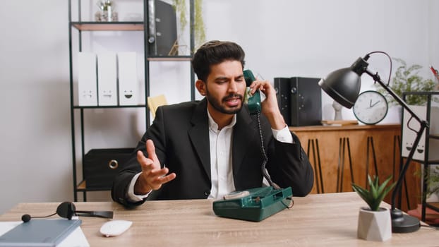 Bearded indian businessman working, having talk with client on retro old-fashioned wired telephone at home office workplace desk. Salesman in formal suit. Remote distant job. Employment occupation