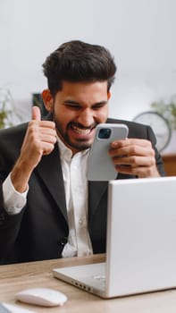 Happy amazed indian businessman working on smartphone shocked by sudden victory game winning lottery jackpot achievemen good career news. Excited bearded hindu man freelancer at home office. Vertical