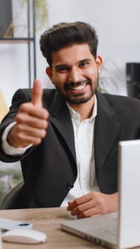 Like. Happy young indian businessman guy working on laptop looking approvingly at camera showing thumbs up, like sign positive something good positive feedback at office. Freelancer man. Vertical shot