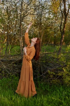 an elegant, sophisticated woman poses standing near a wicker fence in a dacha in a long orange dress, raising her hands up. High quality photo
