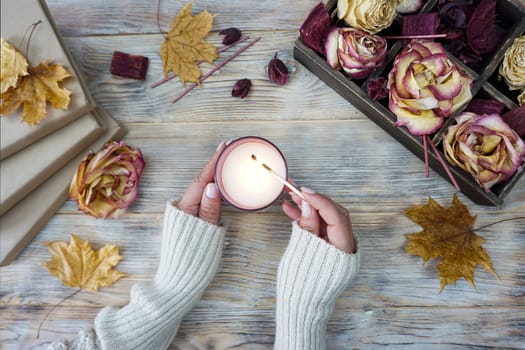 A woman lights an aromatic candle. There are dried roses in a box on the table and yellow maple leaves are lying.