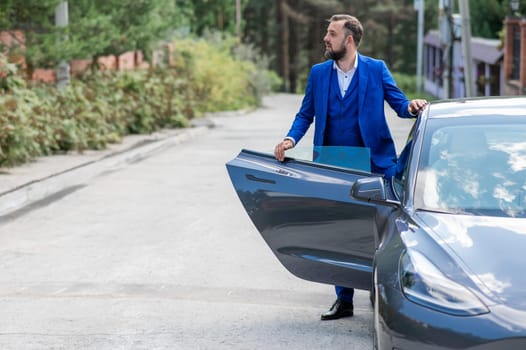 Caucasian bearded man in a blue suit gets out of a black electro car in the countryside in summer