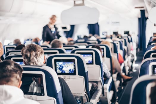 Interior of airplane with passengers on seats and stewardess in uniform walking the aisle, serving people. Commercial economy flight service concept