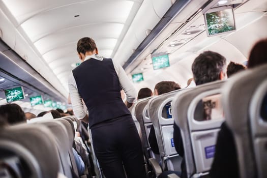 Interior of airplane with passengers on seats and stewardess in uniform walking the aisle, serving people. Commercial economy flight service concept