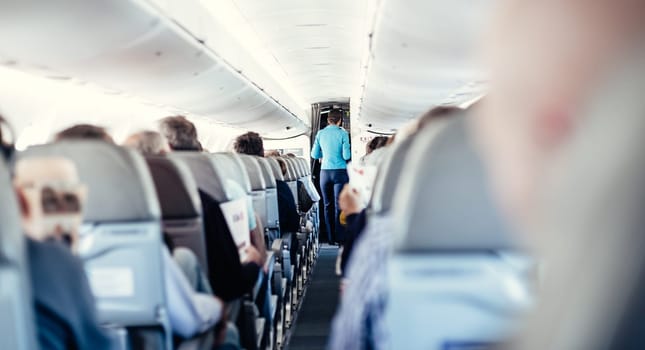 Interior of airplane with passengers on seats and stewardess in uniform walking the aisle, serving people. Commercial economy flight service concept