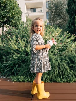 Little girl with a toy hare stands near a flowering juniper bush. High quality photo