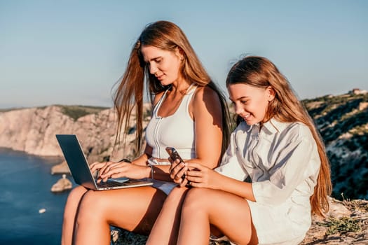 Successful business woman in yellow hat working on laptop by the sea. Pretty lady typing on computer at summer day outdoors. Freelance, travel and holidays concept.