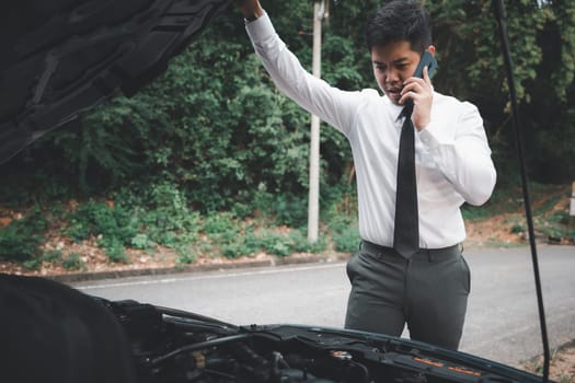 Asian businessman stranded on the road after car breakdown, using phone to call insurance. Candid shot of man in white shirt waiting for assistance and thumbing up, transportation concept.