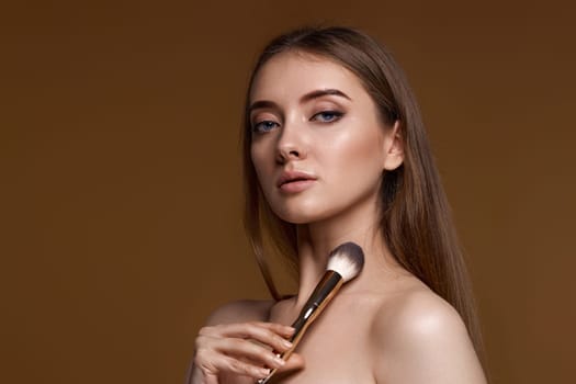 young woman applying blush on cheeks with make-up brush on beige background. copy space