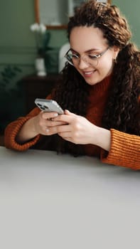 Young smiling woman wearing glasses holding a smartphone using cellphone modern technology, looking at mobile, checking cell phone apps, texting, and browsing the internet for shopping sitting at home