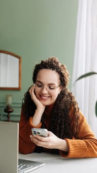 Young happy smiling pretty woman holding smartphone using cellphone modern technology, looking at mobile, checking cell phone apps, watching social media sitting at table at home