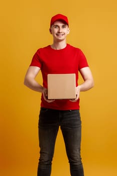 Delivery man employee in red cap, red t-shirt holding cardboard box isolated on yellow background