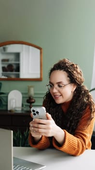Young happy smiling pretty woman sitting at table holding smartphone using cellphone modern technology, looking at mobile phone while remote working or learning, texting messages at home
