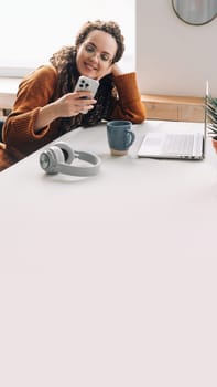 Young happy smiling pretty woman sitting on chair holding smartphone using cell phone mobile technology, looking at mobile phone while remote working or learning, texting messages at home office.