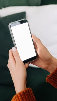 Over shoulder view of woman holding smartphone with white mock up cellular screen in hands using mobile apps on cell phone relaxing on sofa at home. Cellphone display mock up for advertising concept