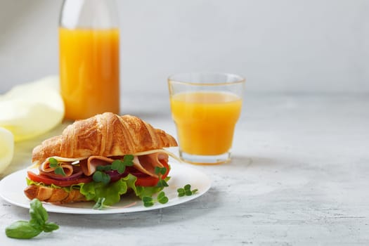 Sandwich with croissant, ham, cheese, tomato, lettuce, microgreens and natural freshly squeezed juice on a gray background.Copy space