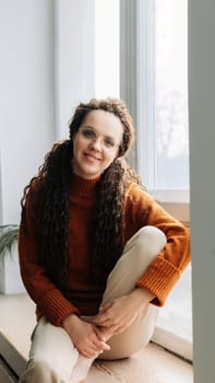 Pretty serene young woman sitting on windowsill relaxing at home looking at camera. Smiling calm lady chilling in apartment, dreaming, thinking of peaceful time enjoying peace of mind. Portrait.