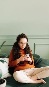 Relaxed young woman sitting on a sofa using a cell phone at home holding a smartphone, looking at cell phones shopping in mobile apps ordering e-commerce products, or checking social media