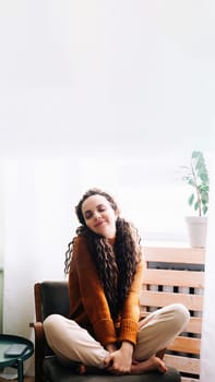 Young relaxed smiling pretty woman relaxing sitting on chair at home. Happy positive beautiful lady feeling joy enjoying wellbeing and lounge chilling near window in modern cozy apartment interior