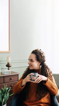 Dreamy young woman savoring warm coffee in cozy home interior. Beautiful lady enjoying a hot drink while relaxing at home. Serene woman with warm mug, lost in thought on cozy winter day.