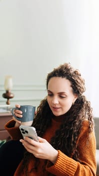 Young woman enjoying a warm drink and online shopping with a smartphone at home. Happy lady with phone and hot beverage, indulging in e-commerce at home. Positive lady enjoying a cozy morning at home.