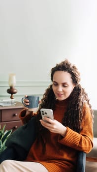 Happy Woman Using Smartphone and Drinking Coffee at Home. Smiling Lady with Phone and Coffee Enjoying E-commerce Shopping. Young Woman Using Mobile Phone and Relaxing with Coffee.