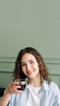 Happy young woman housewife enjoying a cozy break with hot tea on the sofa at home. Smiling lady relaxing with a warm drink and looking at the camera in a close-up indoor portrait