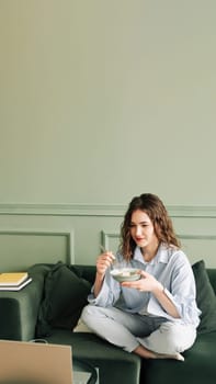Relaxing weekend at home: Young woman in cozy clothes binge-watching TV series, enjoying leisure time with laptop and snacks on the sofa. A young woman who is taking a break from online learning.