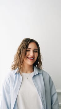 Confident young woman in casual blue tee smiling at camera by window, against white wall. Playful and vibrant indoor portrait