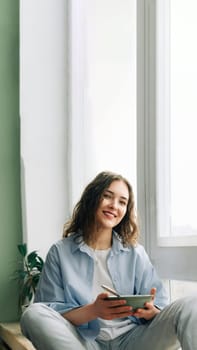 A young woman in jeans and a shirt sitting on the window sill looking through the big window the city holding a bowl of breakfast in her hands, smiling noticing something funny outside.