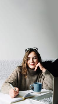 Stylish and focused female PR manager creating content plan for online store, working from home with laptop, notebook and coffee, looking at camera.