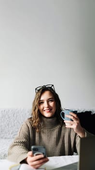 Young student taking a break from online studies, enjoying coffee and phone, laughing at memes and relaxing at kitchen table.