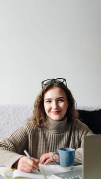 Home office of a young female English tutor with laptop, coffee, and notes, preparing for online lesson with student, sitting at kitchen table. Educator working remotely from home