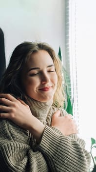 Thoughtful woman standing next to window, looking through, hugging herself, feeling lonely waiting for her boyfriend or dreaming of true love and healthy relationship