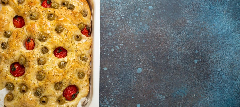 Overhead of Traditional Italian Homemade Flat Bread Focaccia with Green Olives, Olive Oil, Cherry Tomatoes and Rosemary in Baking Tray on Rustic Dark Blue Stone Background, Space for Text