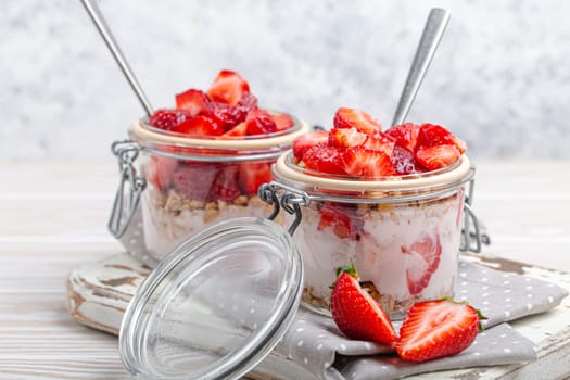 Parfait with Fresh Strawberries, Yoghurt and Crunchy Granola in Transparent Glass Mason Jars on White Rustic Wooden Background from Angle View, Healthy Breakfast or Light Summer Fruit Dessert