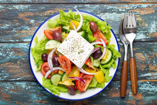 Traditional Greek Salad with Feta Cheese, Tomatoes, Bell Pepper, Cucumbers, Olives, Herbs in white ceramic bowl on blue rustic wooden table background from above, Cuisine of Greece