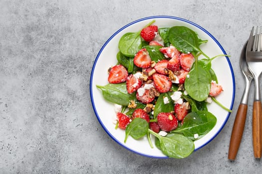 Light Healthy Summer Salad with fresh Strawberries, Spinach, Cream Cheese and Walnuts on White Ceramic Plate, Grey rustic stone Background Top View Copy Space.